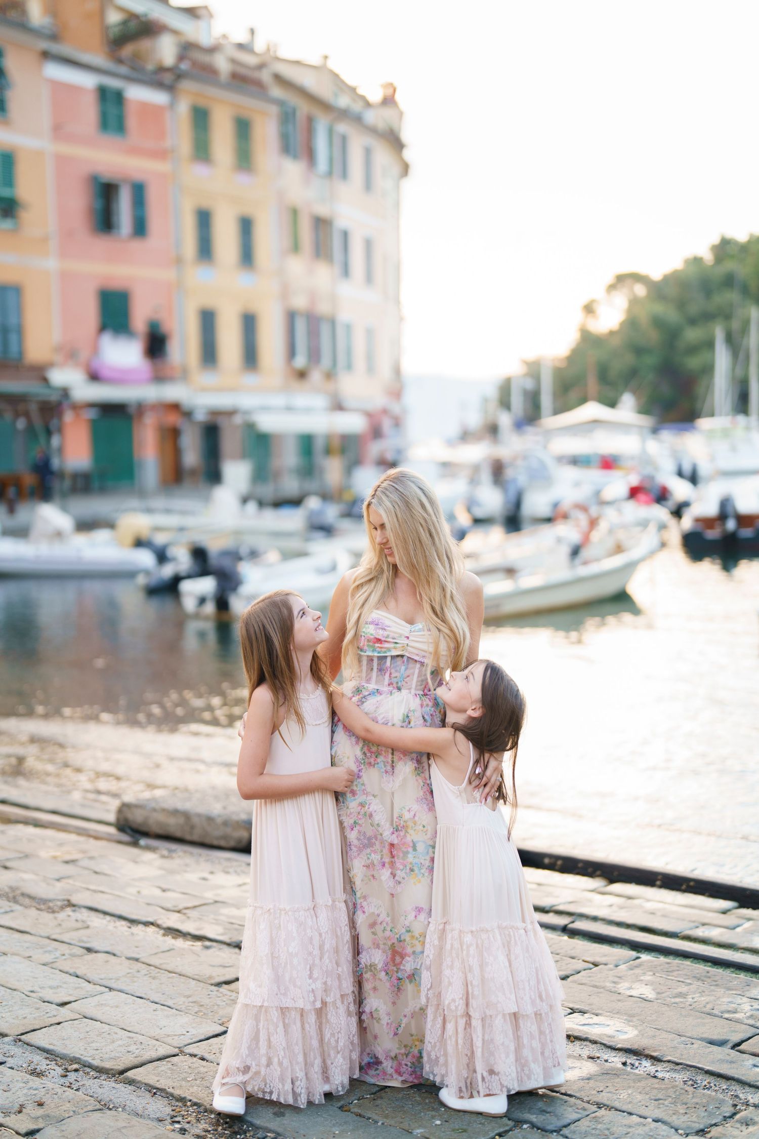 Two people in floral dresses share a sweet moment on a cobblestone street near boats in an Italian coastal town.