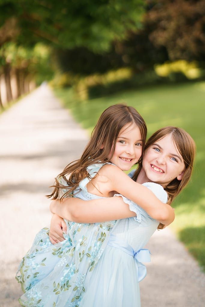 Two young girls in blue dresses embrace and smile while standing on a tree-lined path in a garden setting.