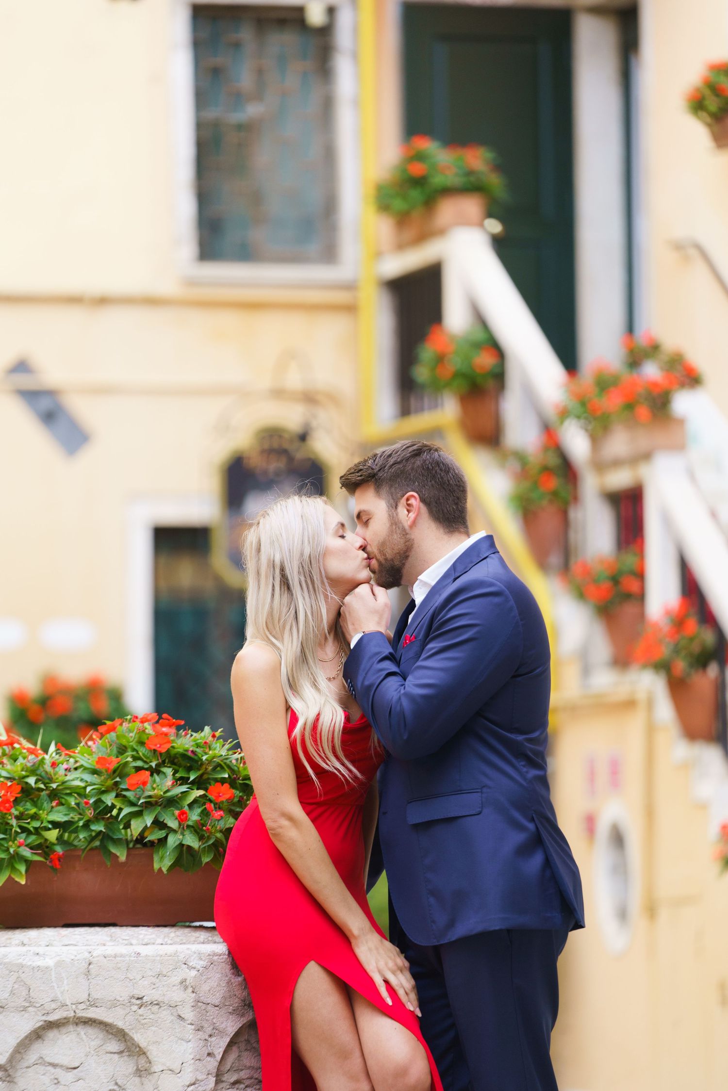 Romantic couple embraces on Italian street with red flowers and architecture in background.