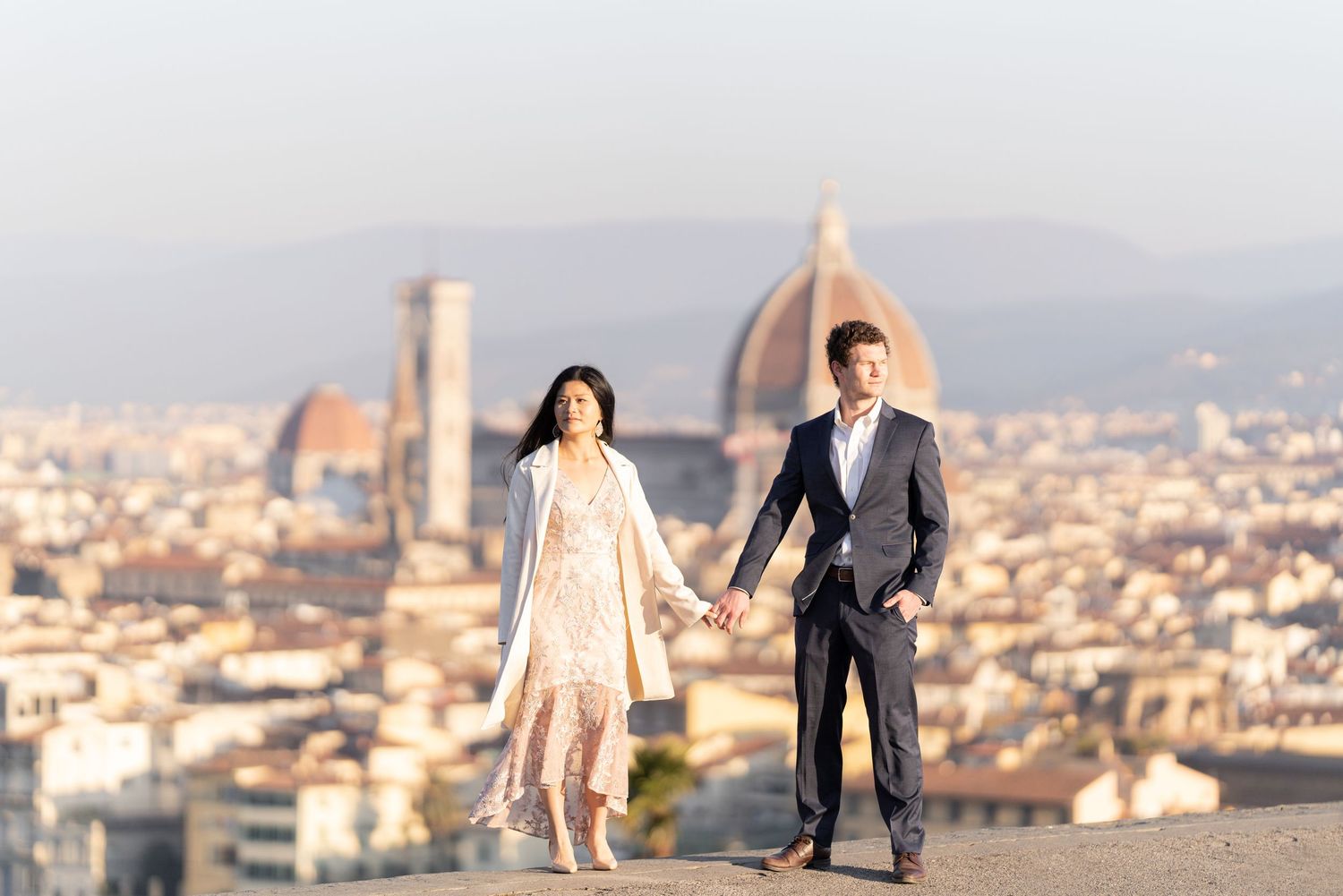 A couple stands holding hands overlooking Florence, Italy with the iconic Duomo cathedral dome visible in the cityscape.