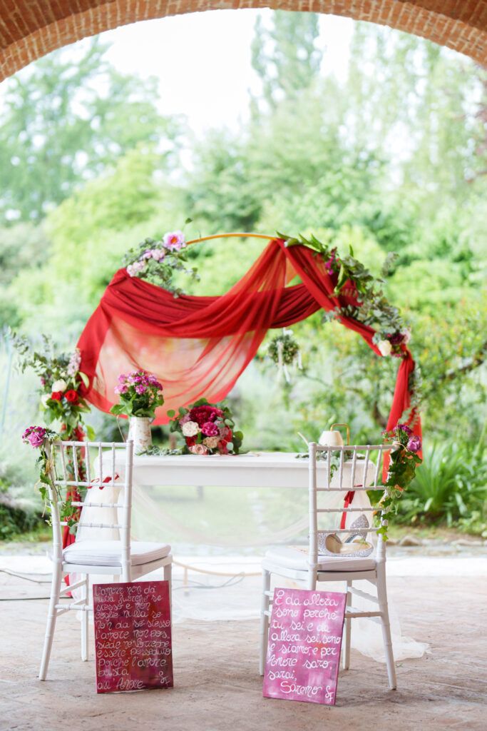 Wedding ceremony setup with red fabric draping and floral arrangements on white chairs.