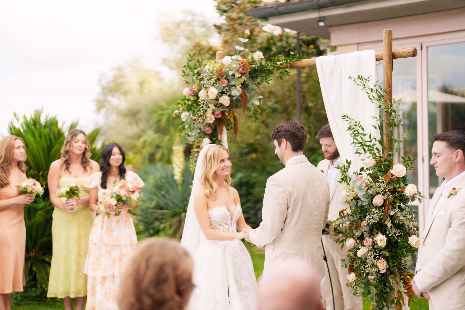 An outdoor wedding ceremony under a floral arch with bridesmaids in pastel dresses.