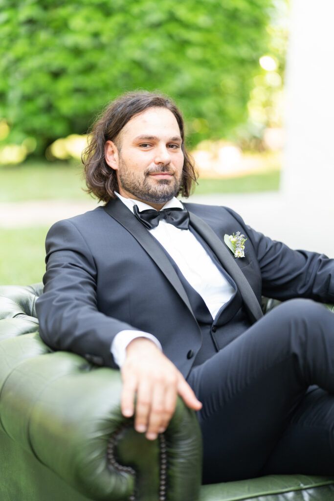 Formal portrait of groom in black tuxedo relaxing on leather chair outdoors.