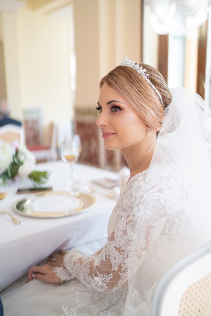 Bride in lace gown seated at elegant reception table with ornate place settings.