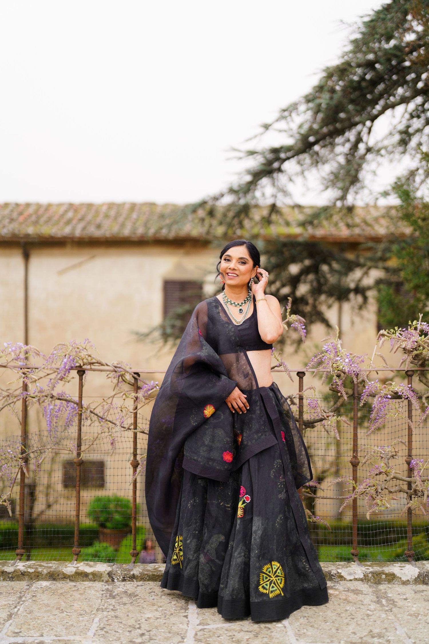 Person in a black dress sitting on stone wall with flowering trees and rustic buildings in background.