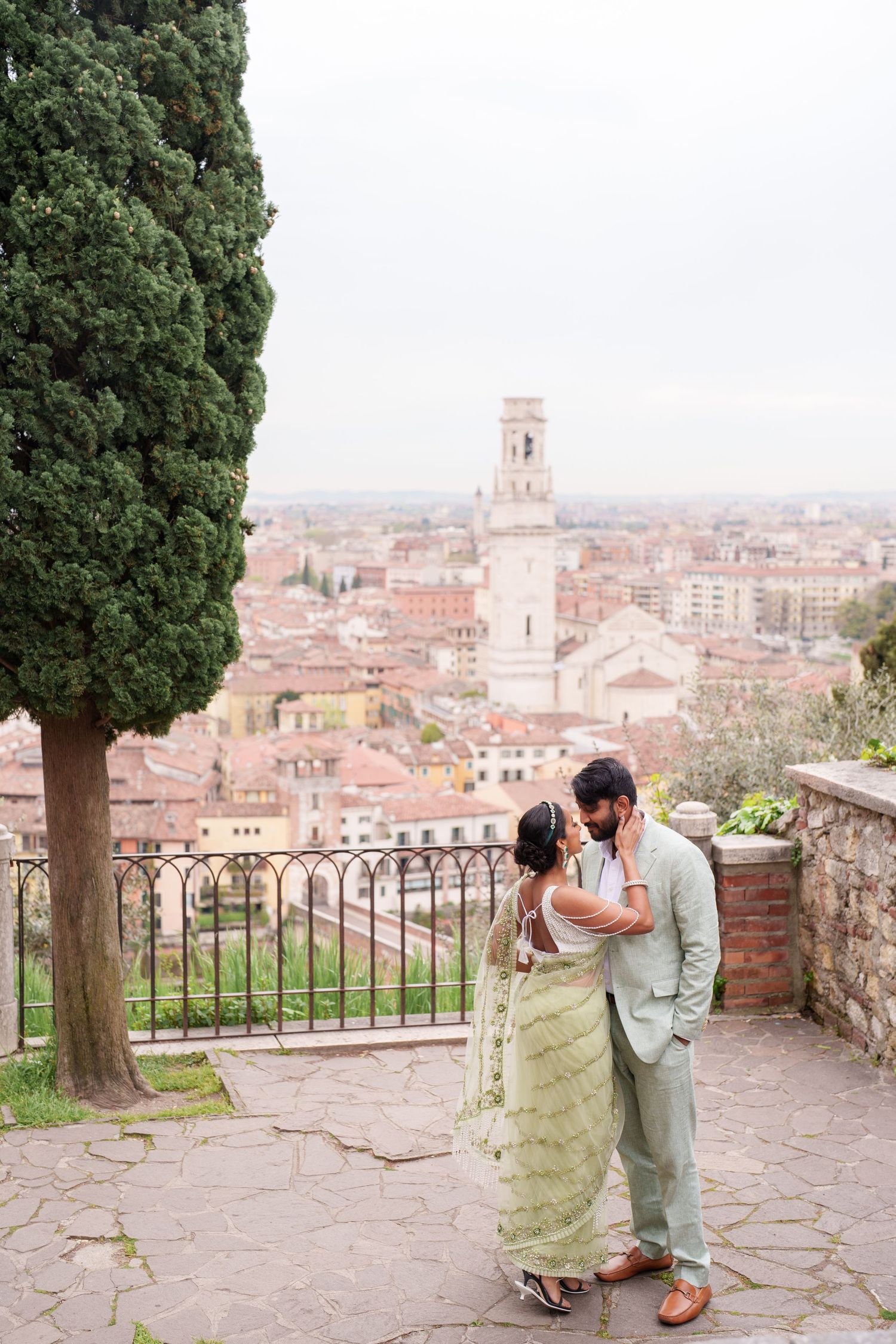 Couple embraces on stone terrace overlooking historic European cityscape with red rooftops and church tower in background.