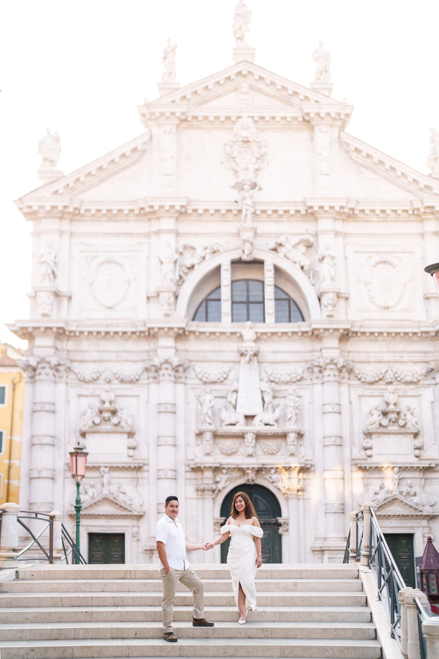 Couple poses joyfully in front of an ornate white baroque church facade with arched windows and decorative details.