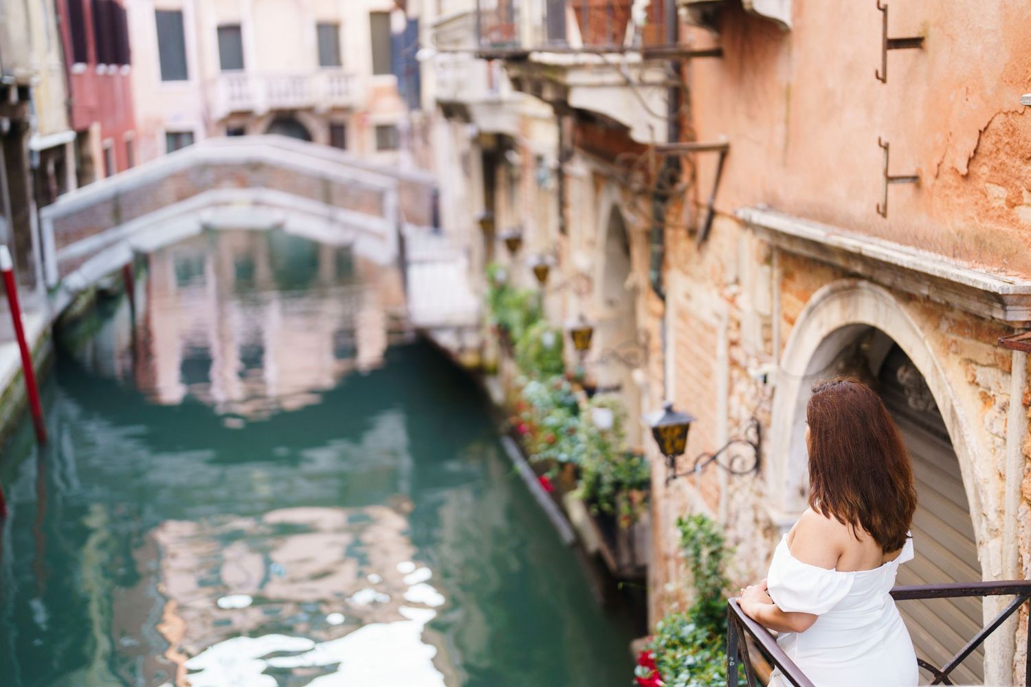 A narrow canal in Venice with a stone bridge and pink buildings reflecting in the turquoise water.