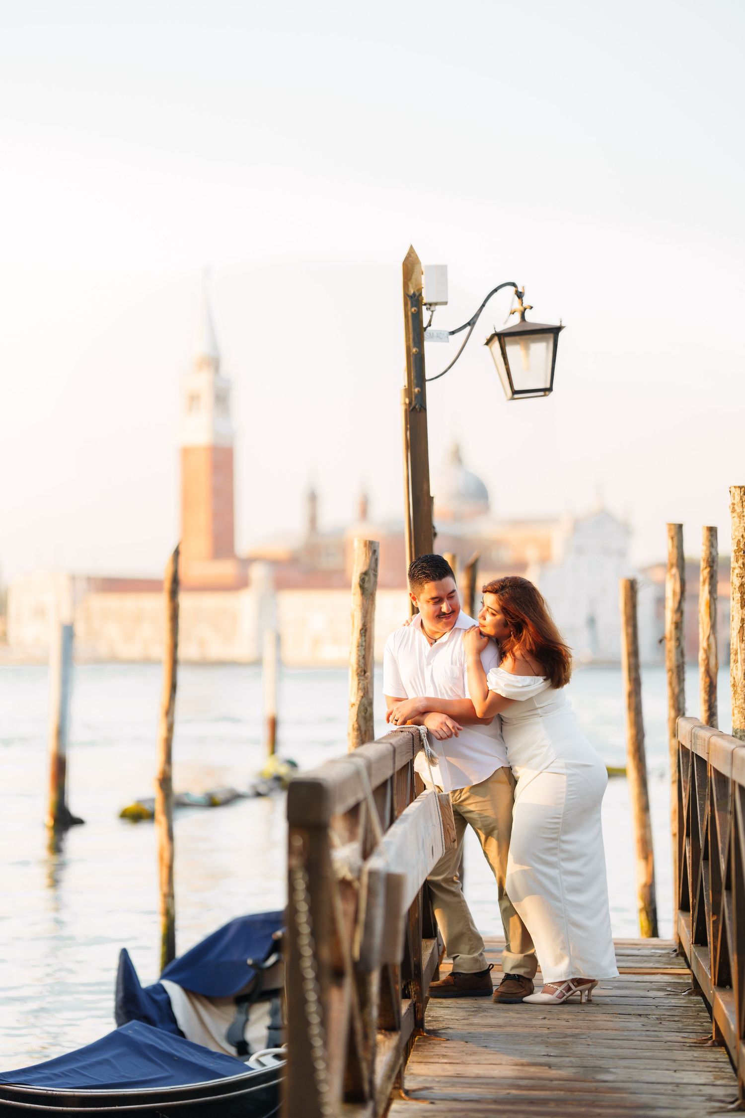 Romantic couple embraces on a wooden pier overlooking Venice's iconic waterfront with gondolas and buildings at sunset.