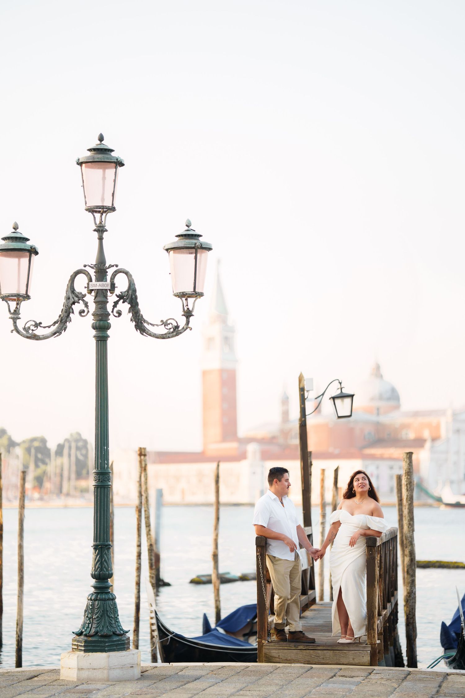 Romantic couple enjoying sunset views by ornate lamp posts along Venice's iconic waterfront with gondolas in background.