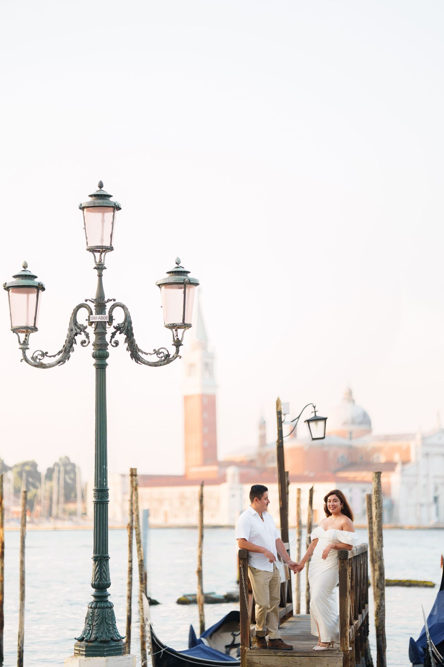 Romantic couple standing by ornate street lamp and gondolas along Venice canal at sunset with church dome in background.