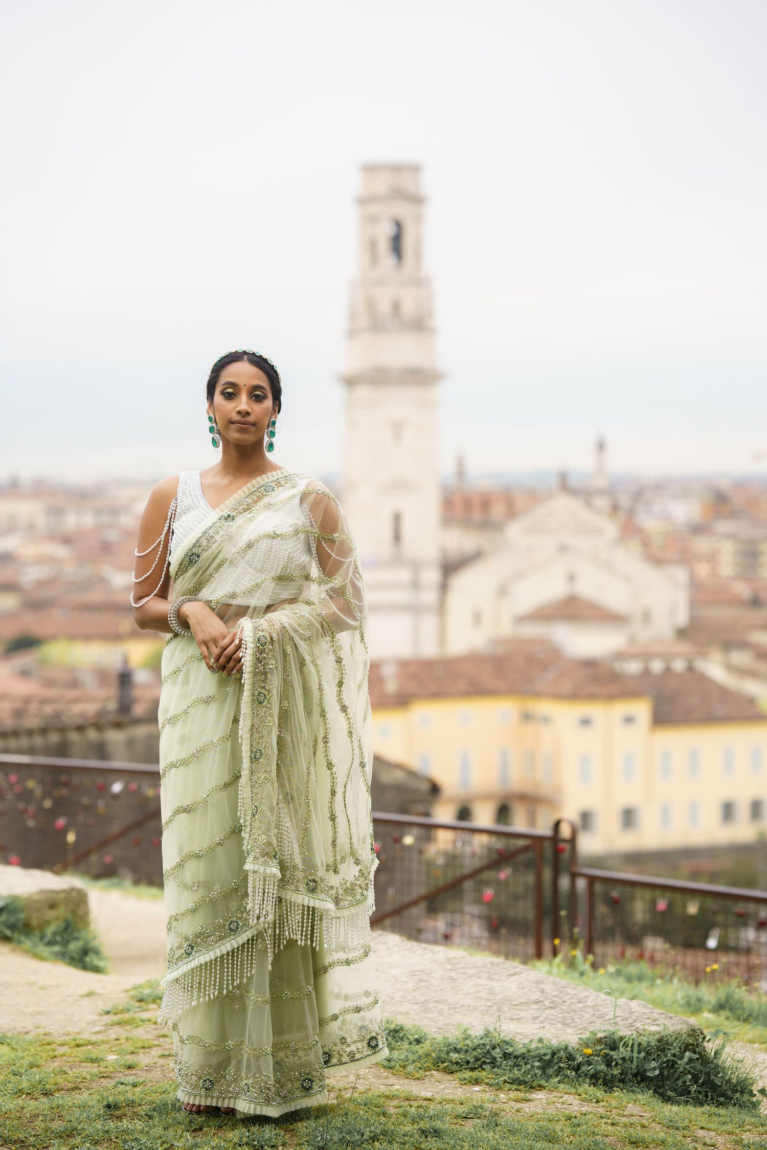 A person in a light green traditional sari poses against a scenic Italian cityscape with historic architecture.