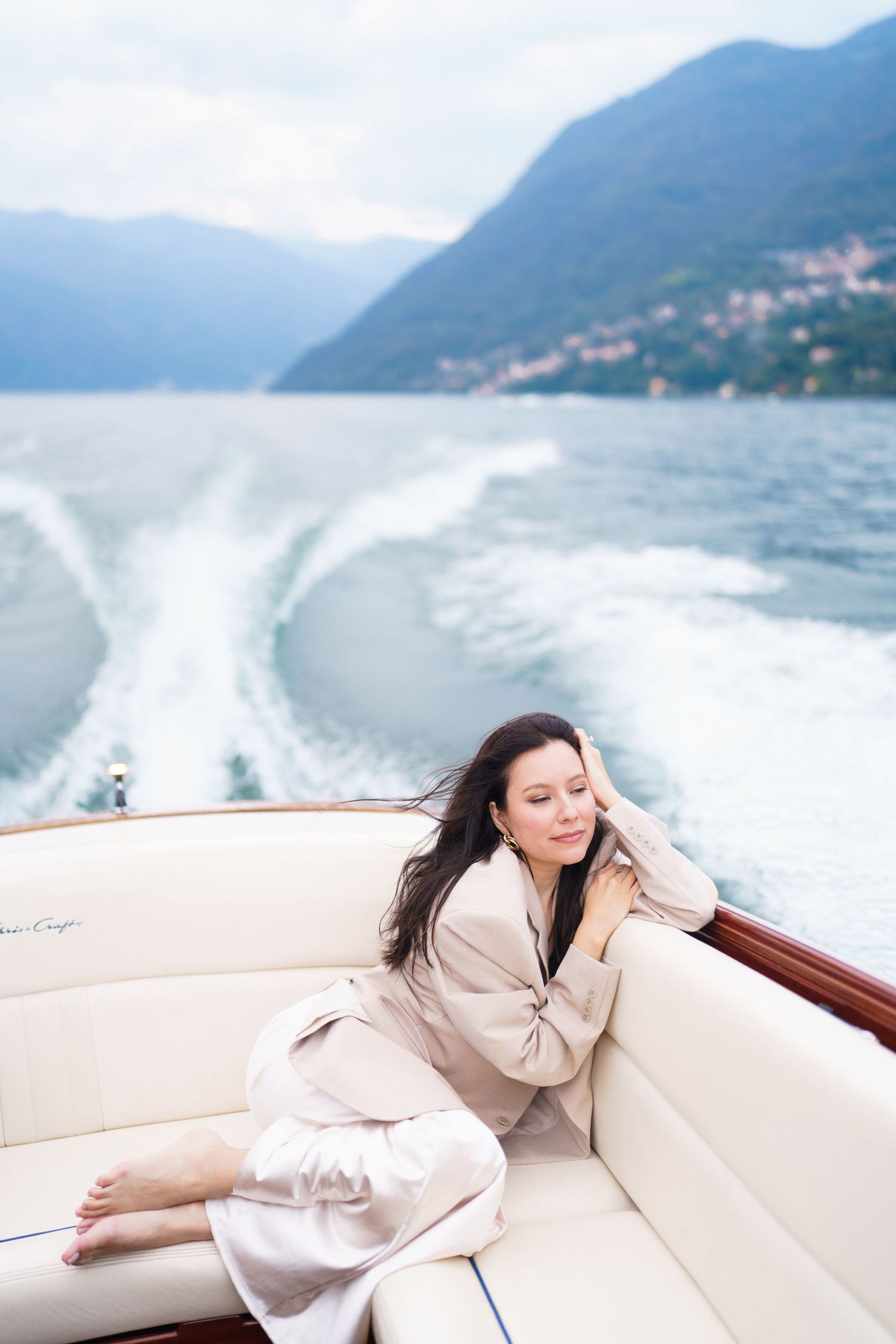 Woman in beige loungewear relaxes on white boat cushions with scenic mountain lake views in background.