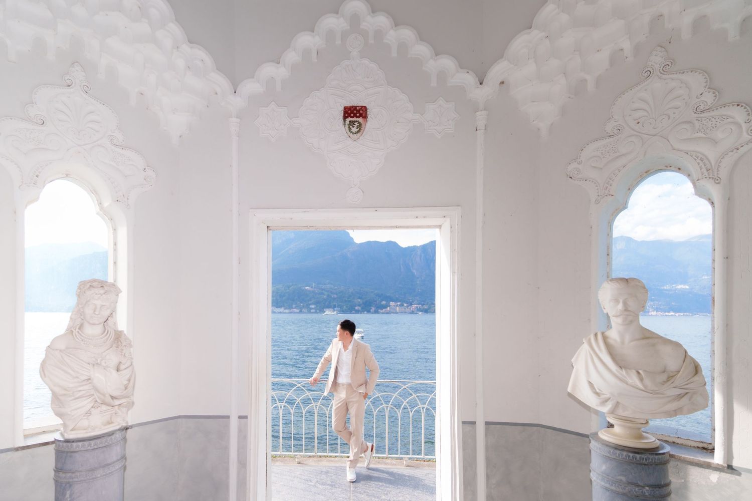 A person stands in an ornate white archway overlooking a coastal view with marble busts on either side.