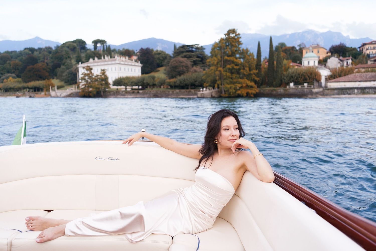 A person in a white dress relaxes on a wooden boat deck with coastal villas and trees visible along the Mediterranean shore.