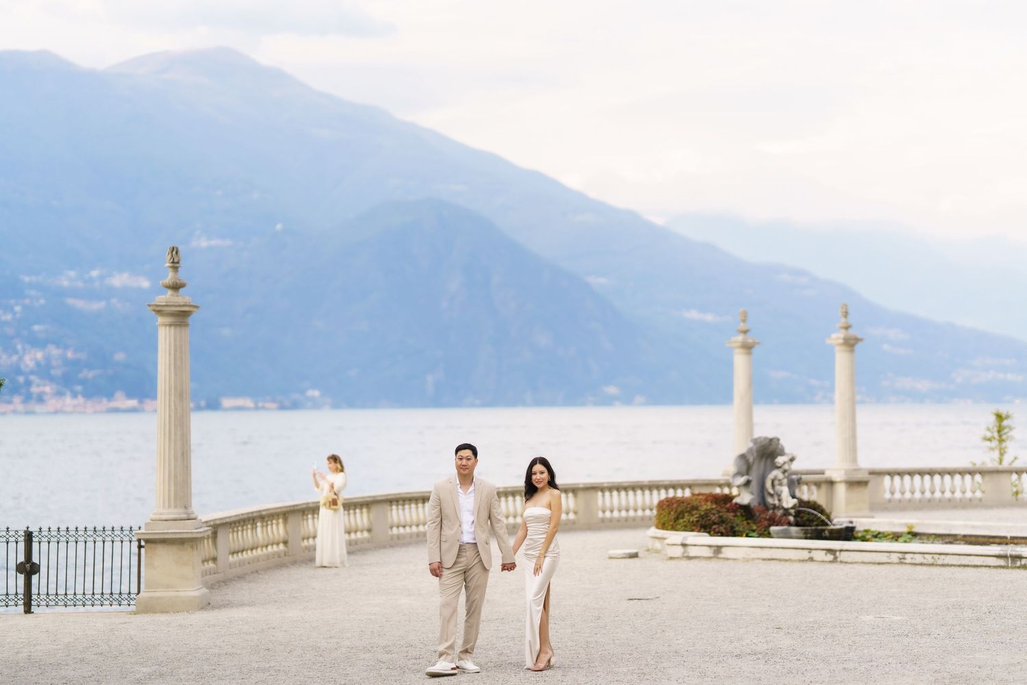 Couple walking along elegant lakeside terrace with stone columns and mountain backdrop in Lake Como, Italy.