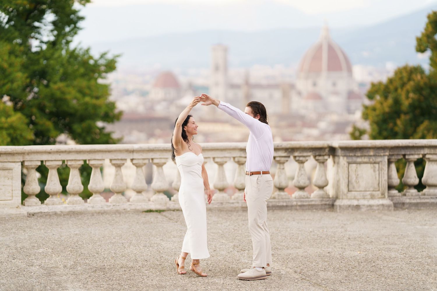 A romantic couple in white outfits walking along a scenic terrace overlooking Florence, Italy during golden hour.