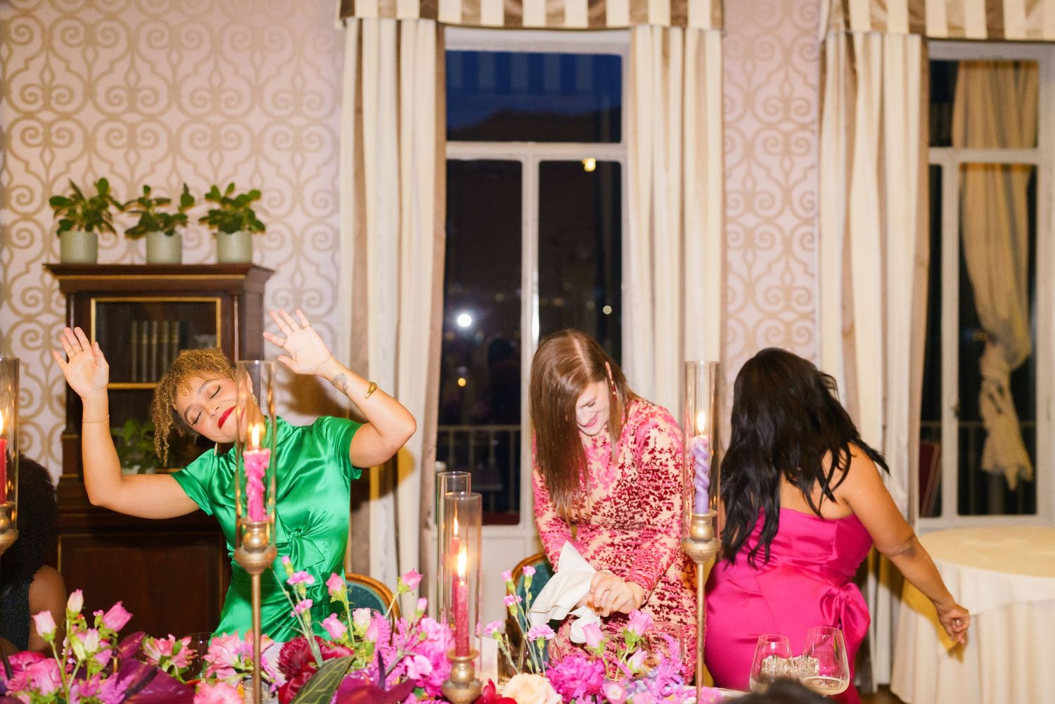 Three women in elegant attire laugh and celebrate at a candlelit dinner table adorned with pink floral arrangements.