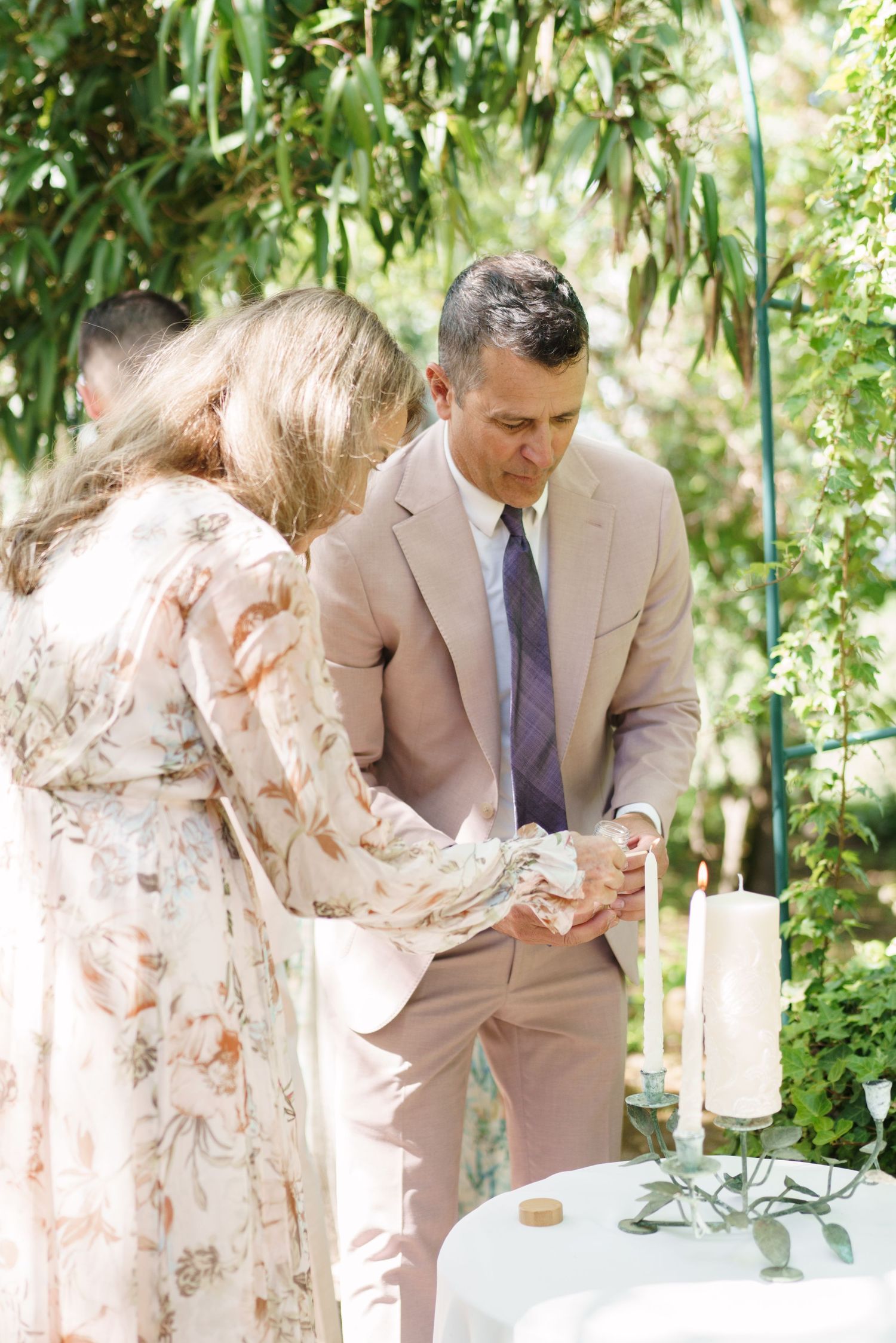 Man in tan suit and woman in floral dress lighting unity candles at an outdoor wedding ceremony.