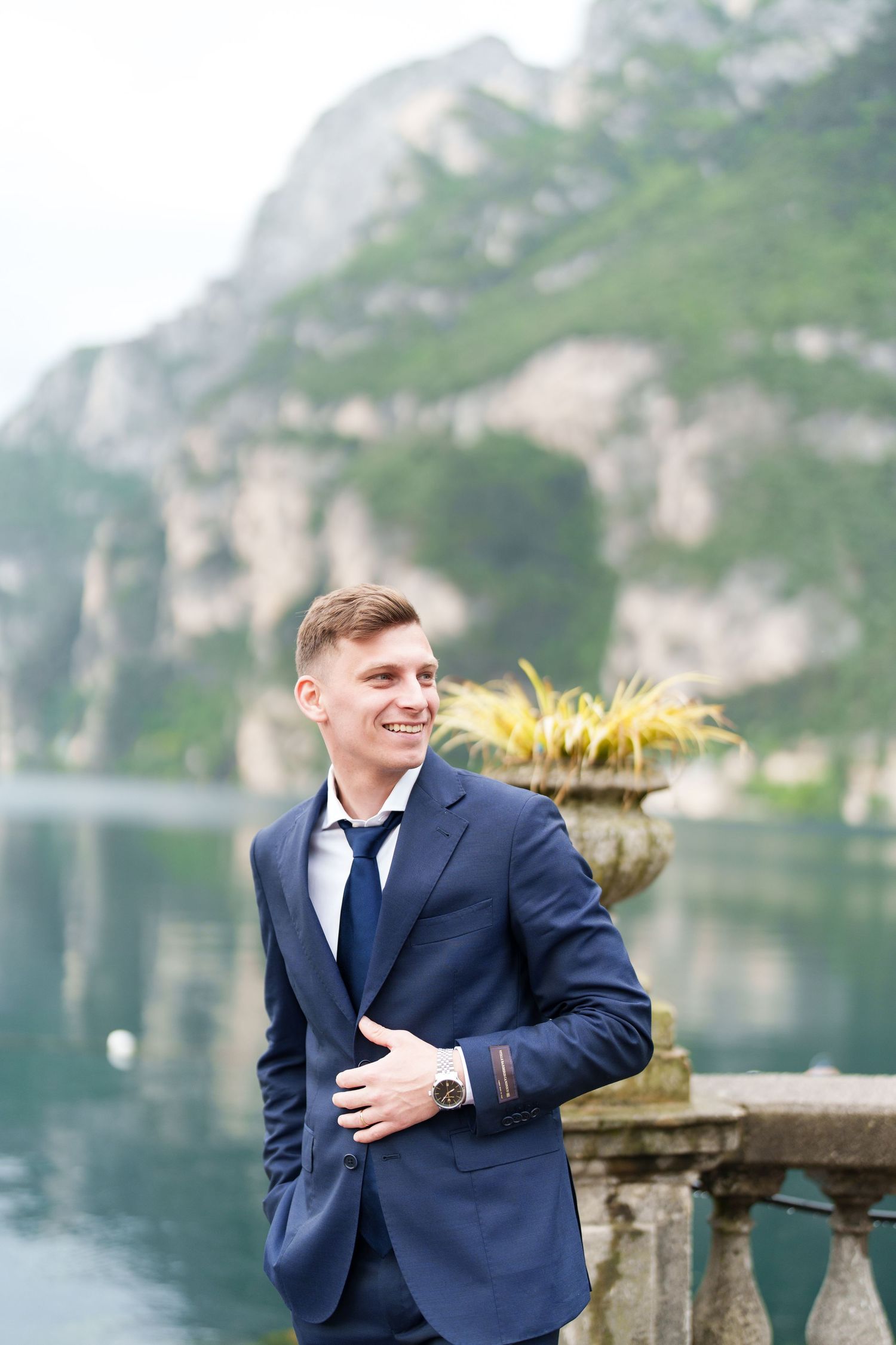 A person in a navy suit stands by an ornate stone balustrade overlooking a misty mountain lake.