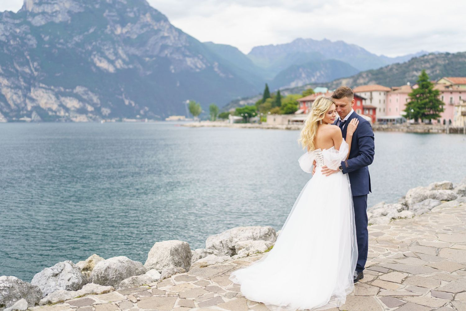 A couple in wedding attire embraces on a stone path along a scenic mountain lake in Italy.