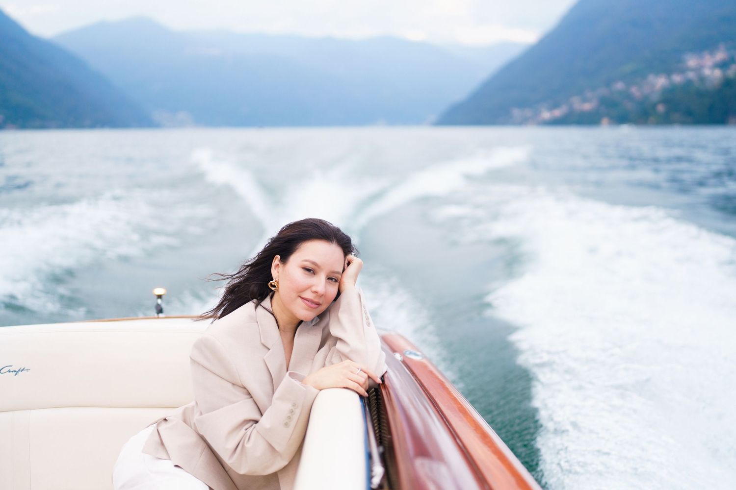 Woman in beige outfit enjoys a scenic boat ride across a misty mountain lake surrounded by dramatic peaks.