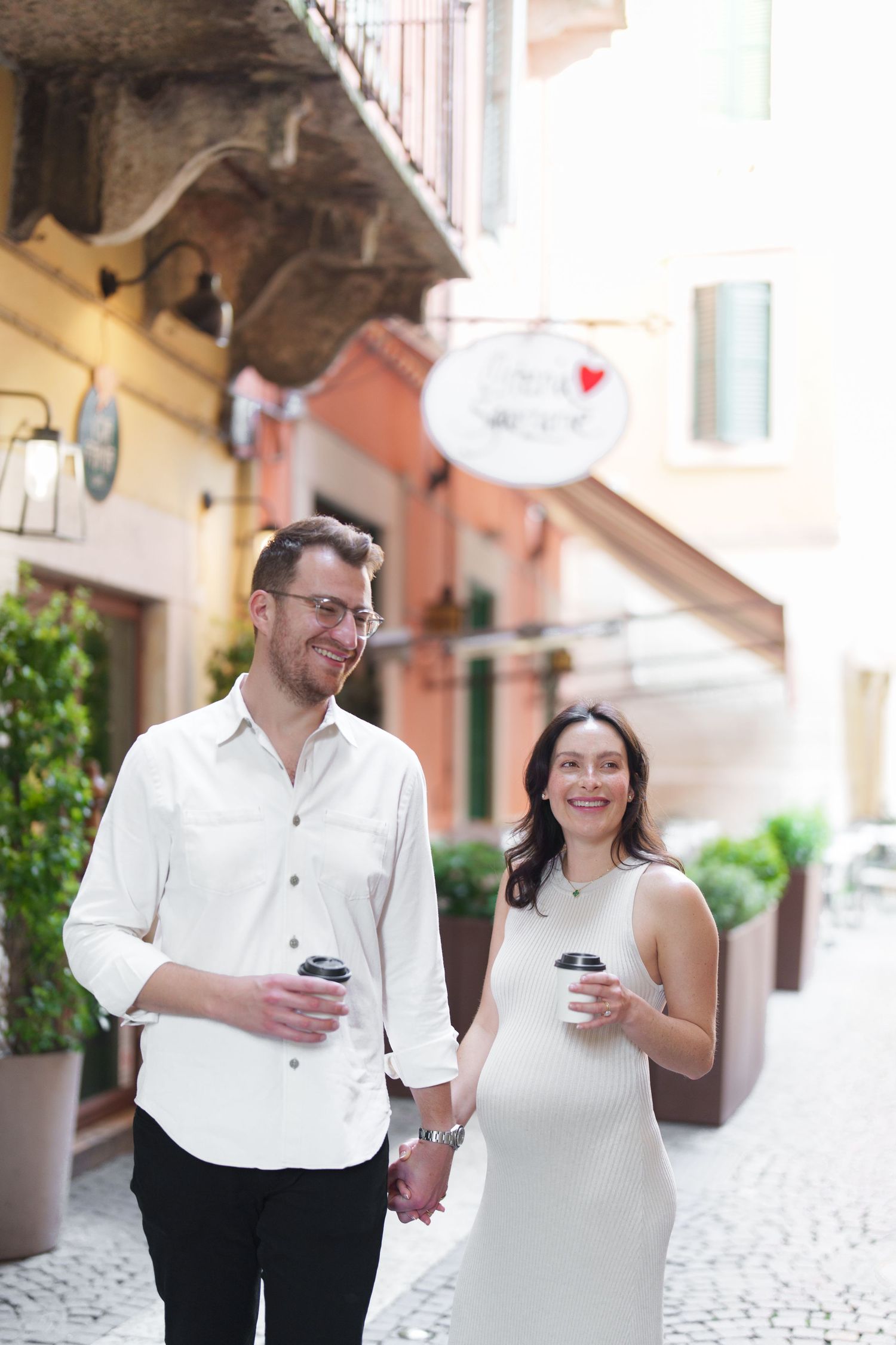 A couple in white outfits walks together holding coffee cups while strolling through a charming European alleyway.