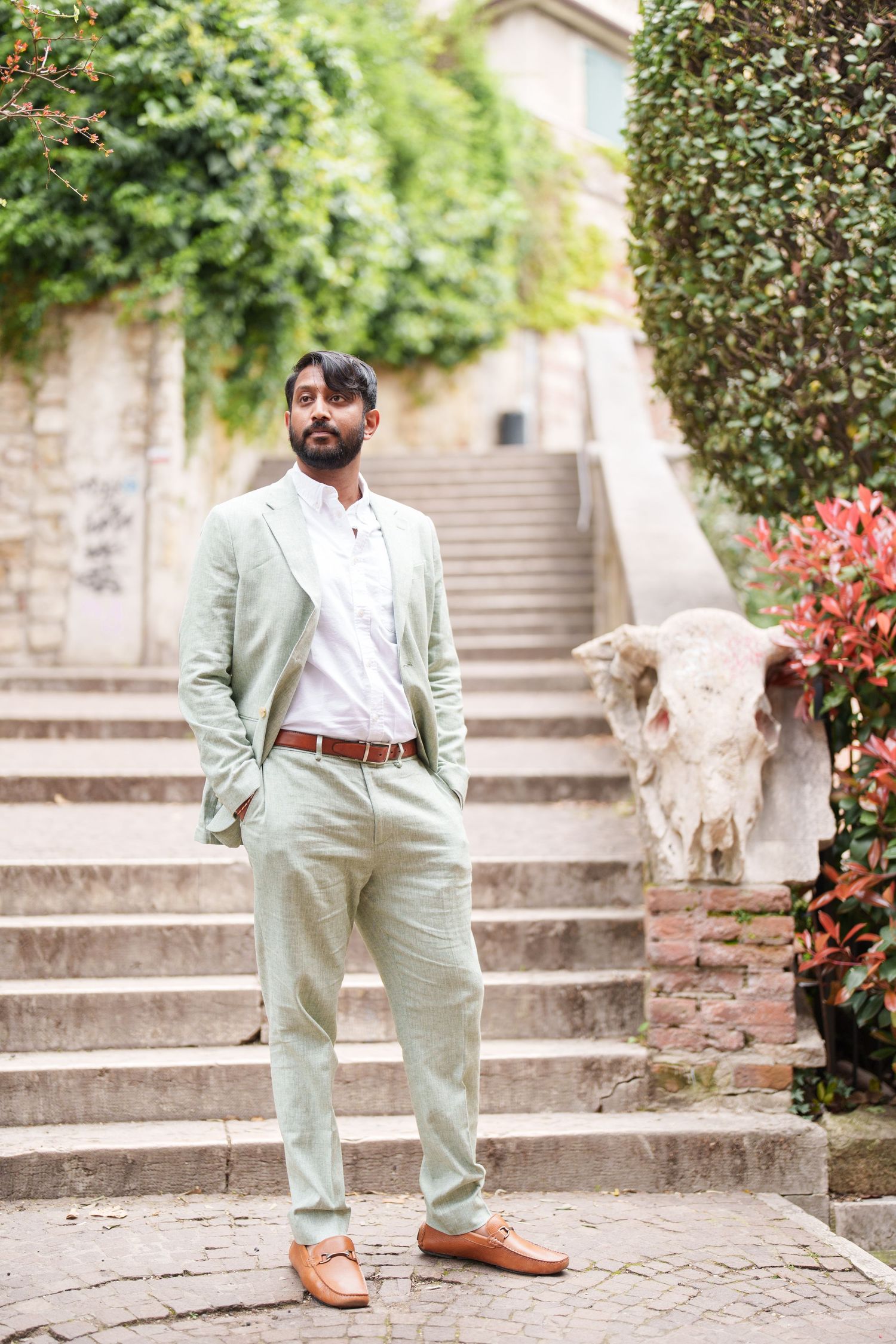 photographer verona A person in a mint green suit and brown shoes poses on stone steps surrounded by lush garden landscaping.