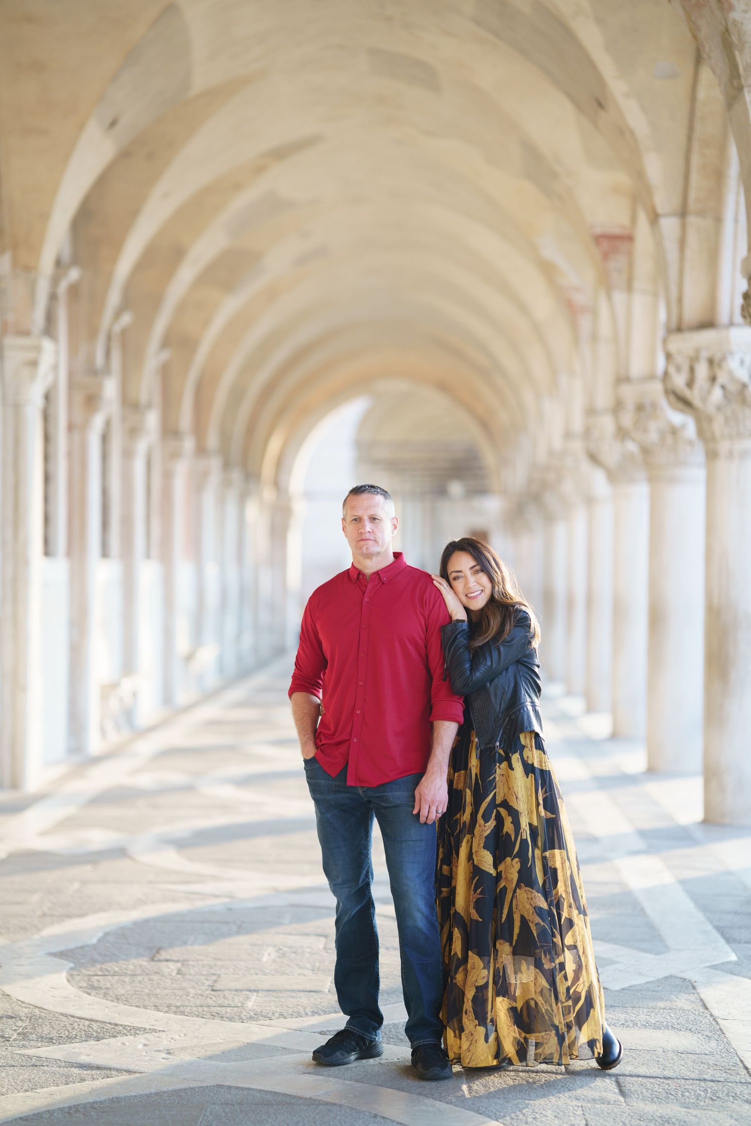 Couple poses in elegant arched corridor with repeating columns and natural lighting creating a romantic atmosphere.