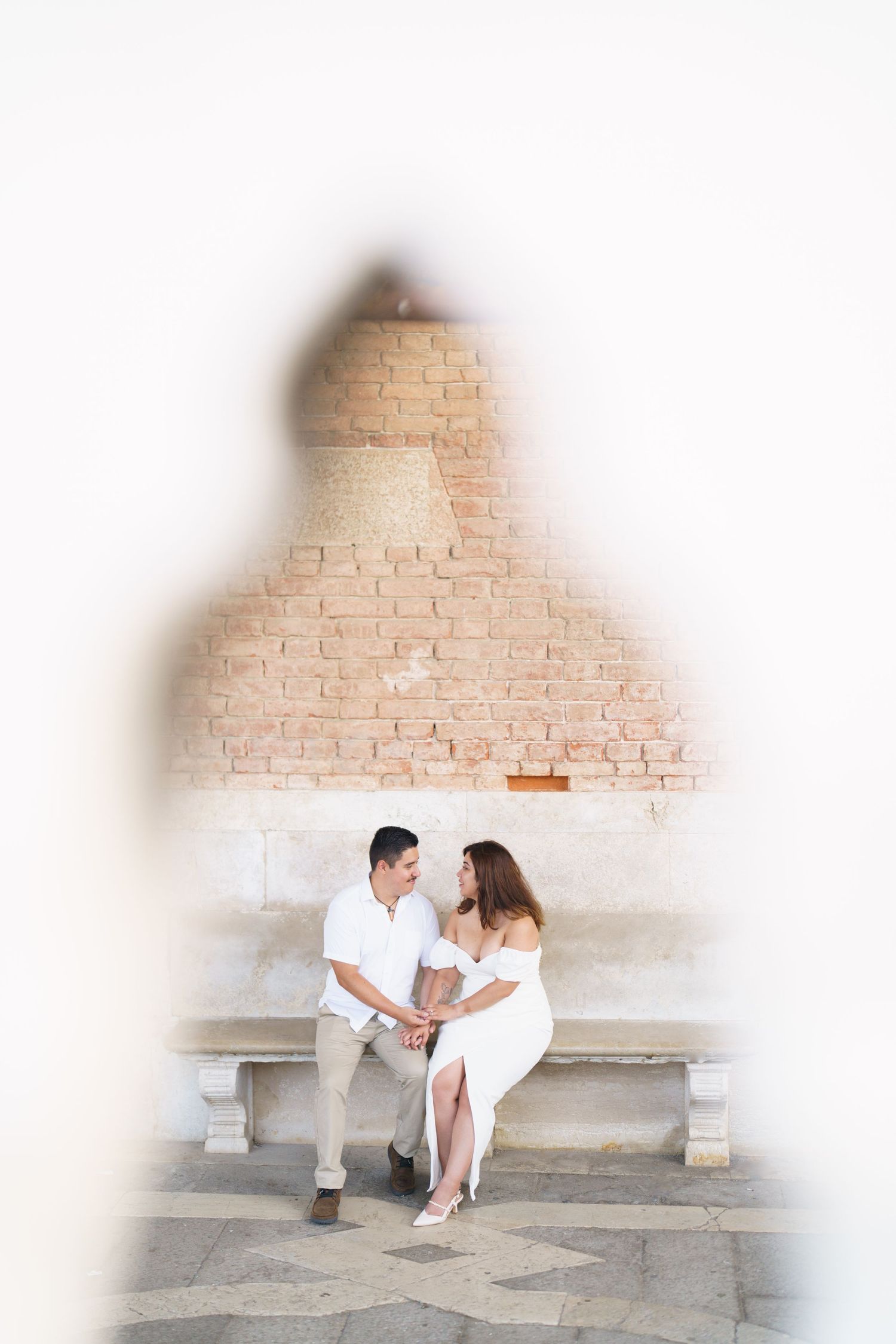 A romantic couple in white outfits sits together on stone steps against a rustic brick wall backdrop.