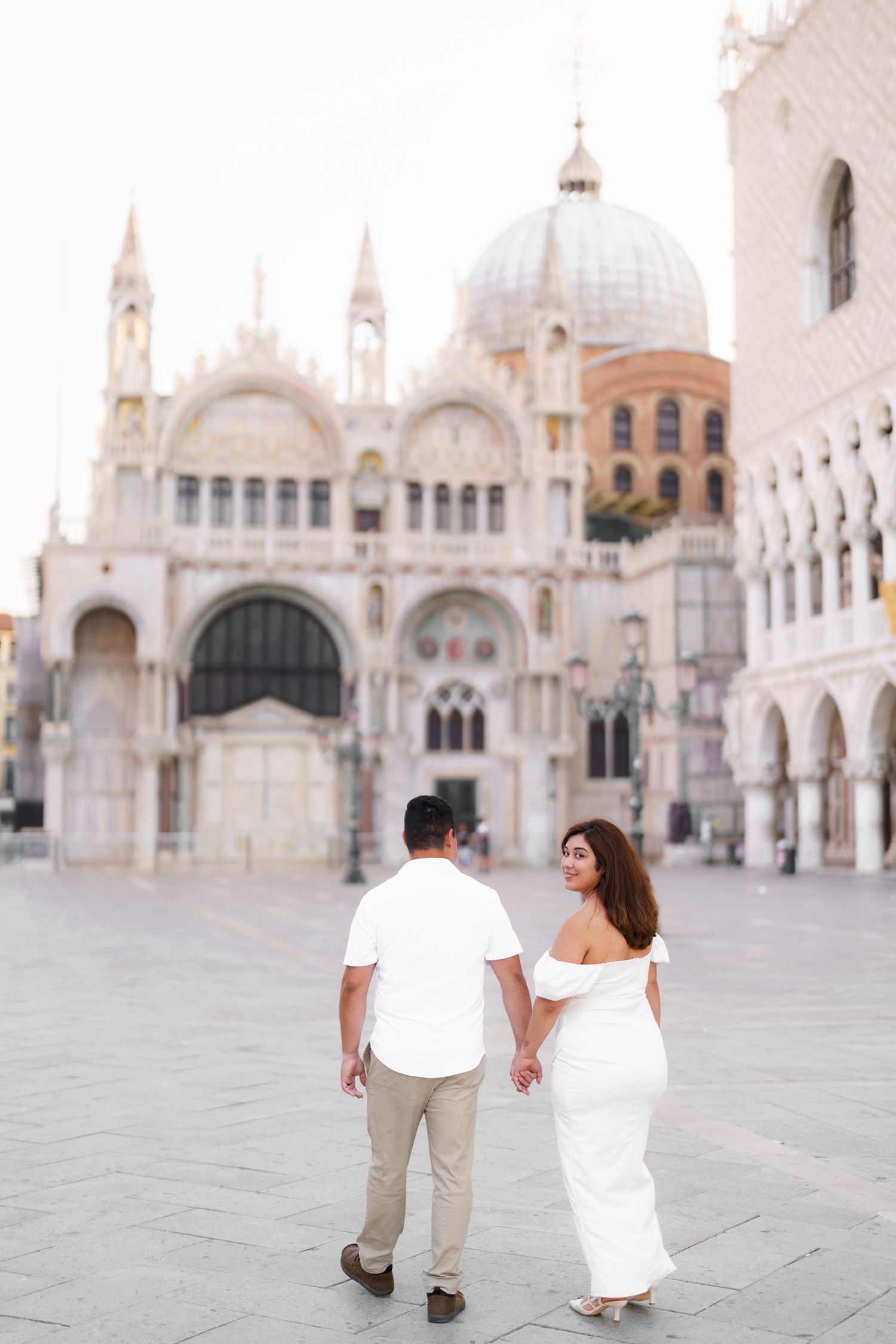 A couple poses in white and beige outfits at Saint Mark's Square in Venice with the basilica in the background.