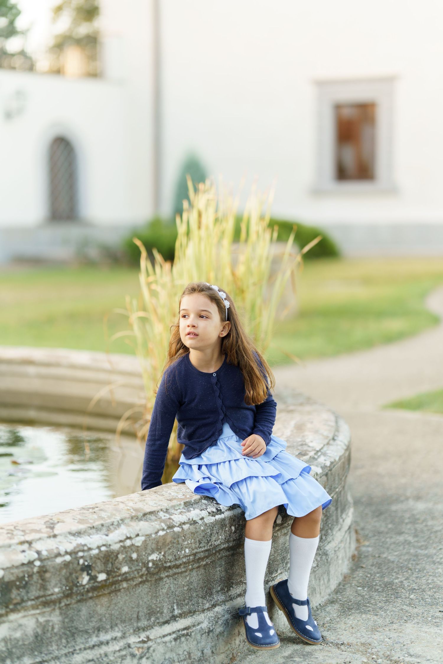 Child in blue dress and white socks sits on stone ledge near water in outdoor setting.