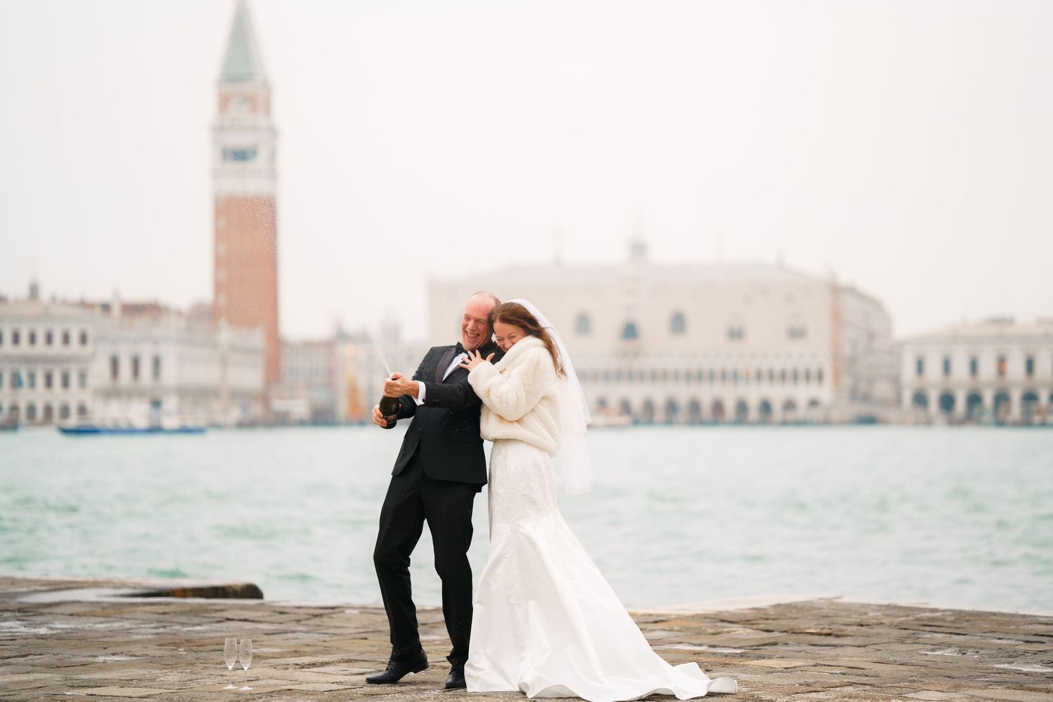 A couple in wedding attire share a romantic moment on a pier overlooking Venice's St. Mark's Square and bell tower.