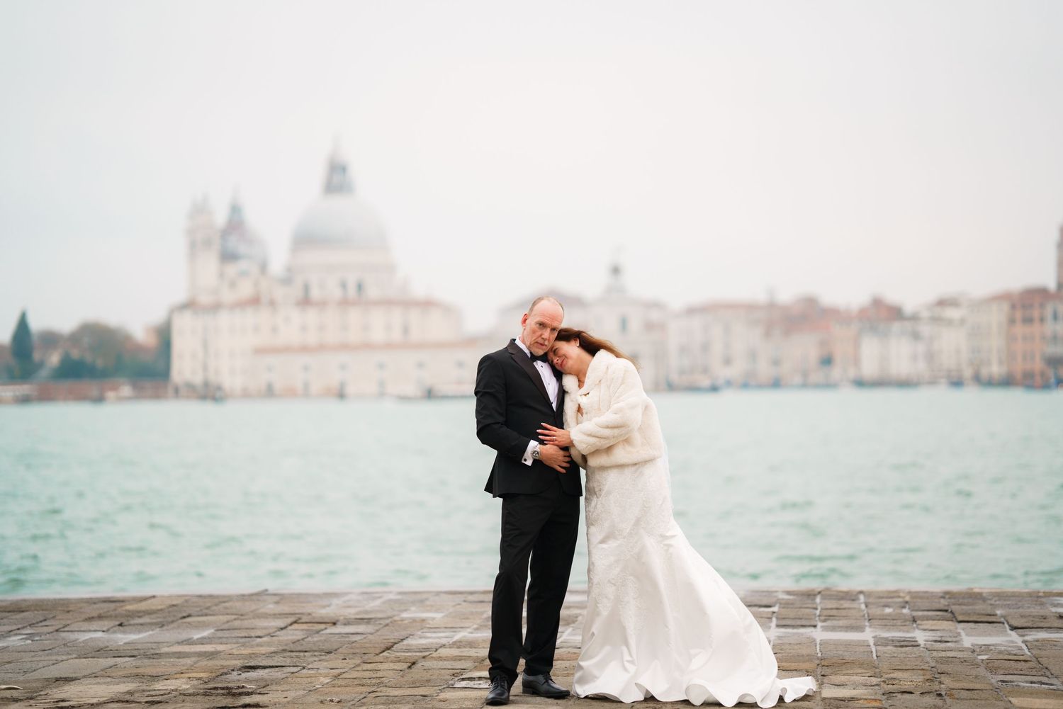 A couple in wedding attire embrace on a pier overlooking Venice's Santa Maria della Salute church and lagoon.