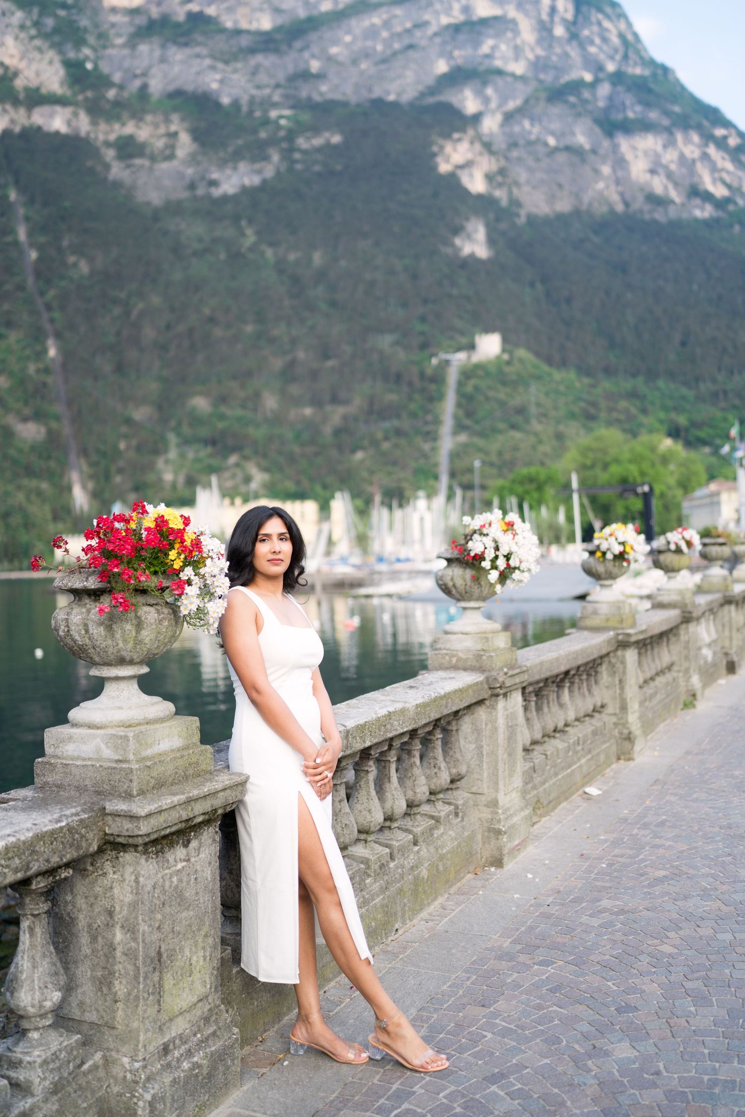 Elegant white dress against mountain lake backdrop with stone balustrade and floral arrangements in Lake Como, Italy.
