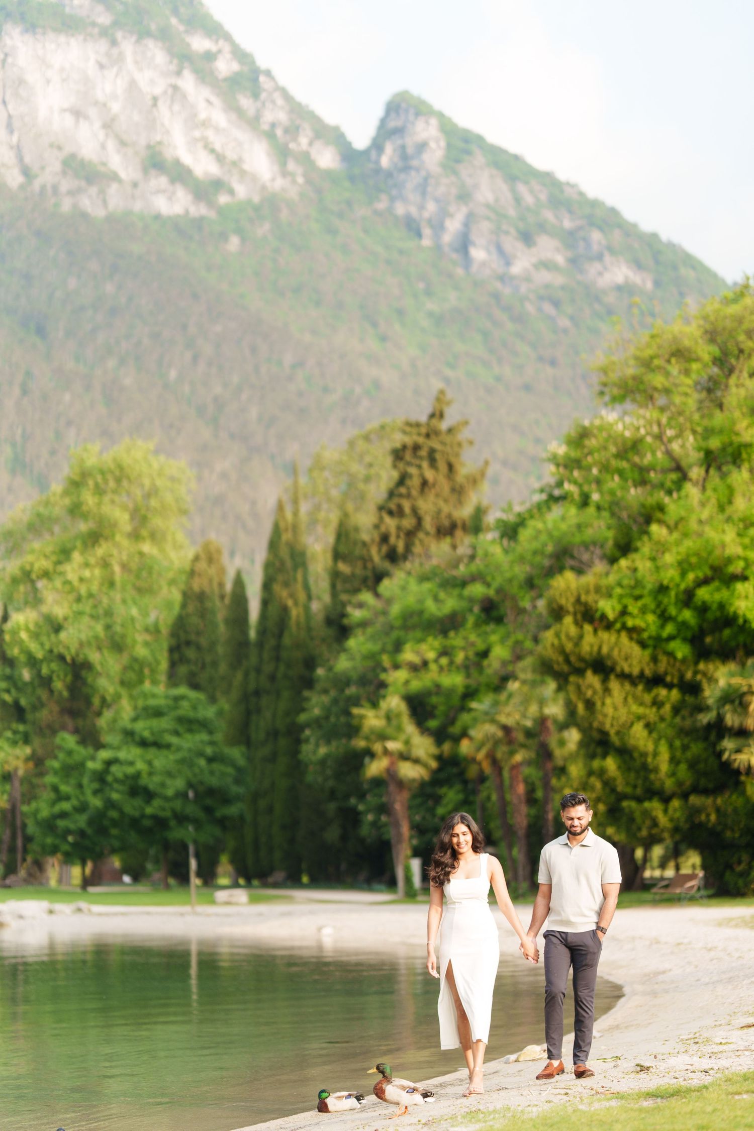 Couple walking and sitting along peaceful mountain lakeside beach surrounded by lush green trees and mountains.