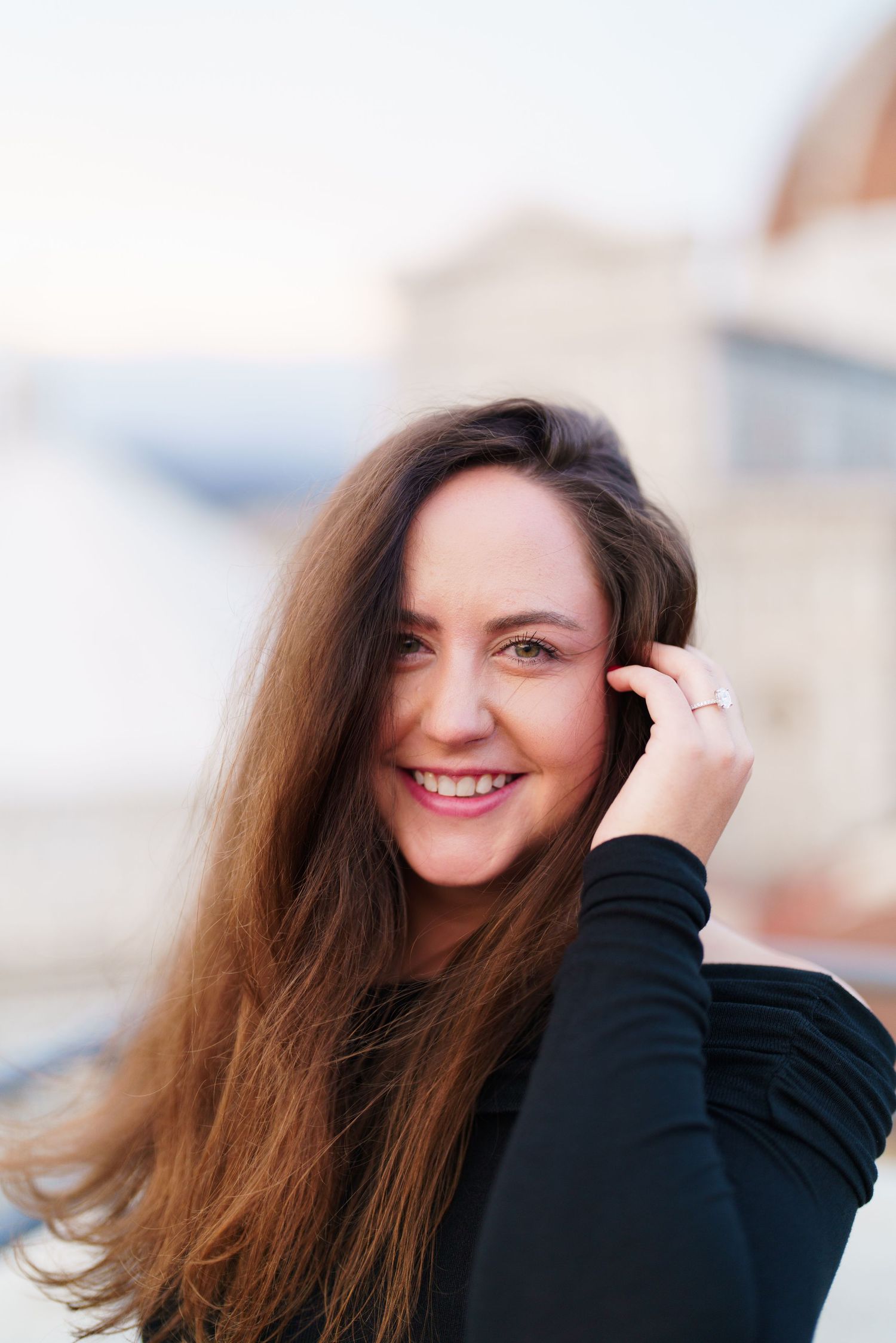 A person with long brown hair smiles while wearing a black sweater against a blurred background.