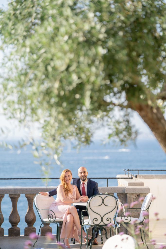 Mediterranean seaside terrace with chairs and tables under a tree canopy.