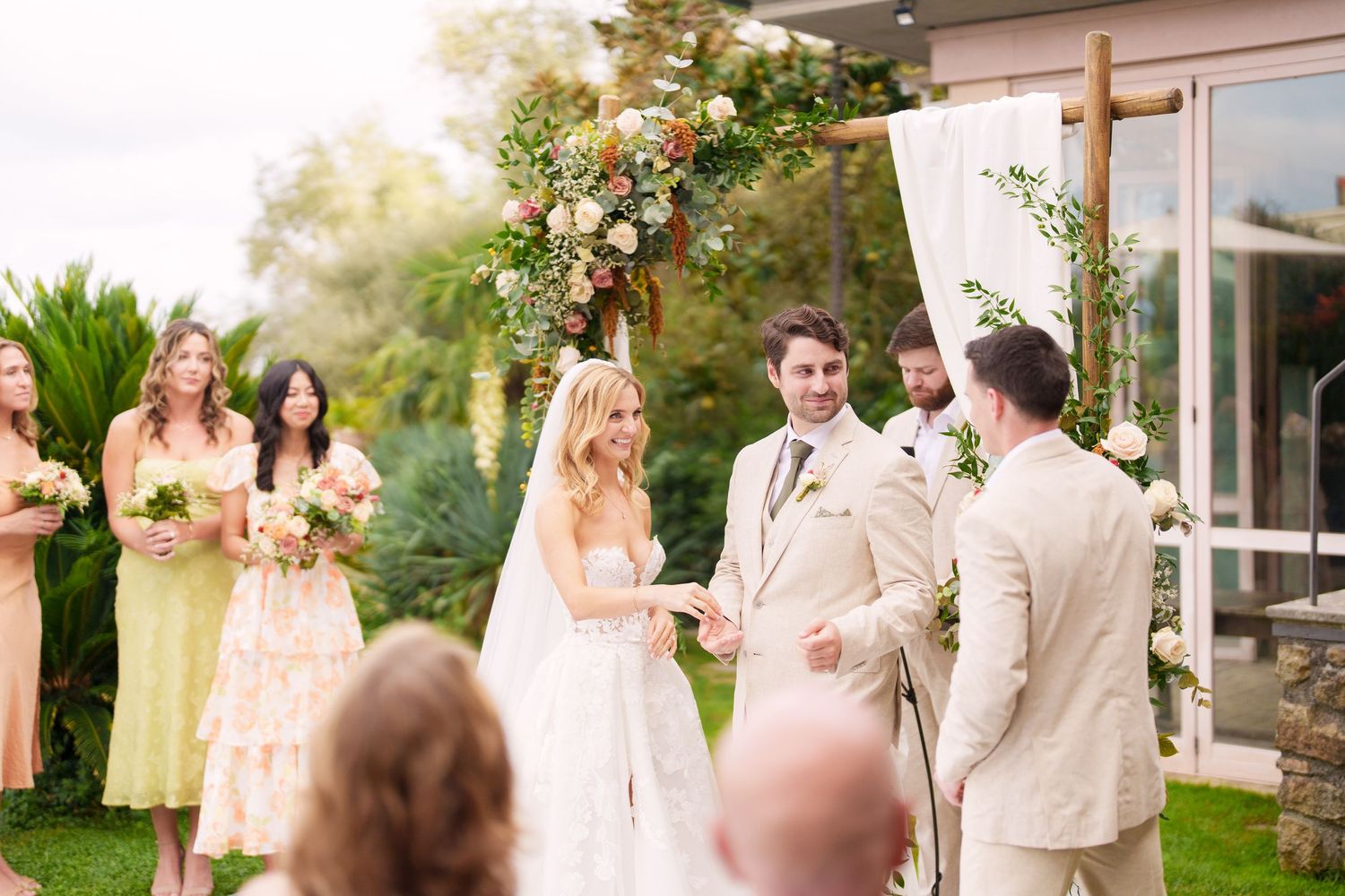 Wedding party gathers under a floral arch for an outdoor ceremony with pink and white decorations.