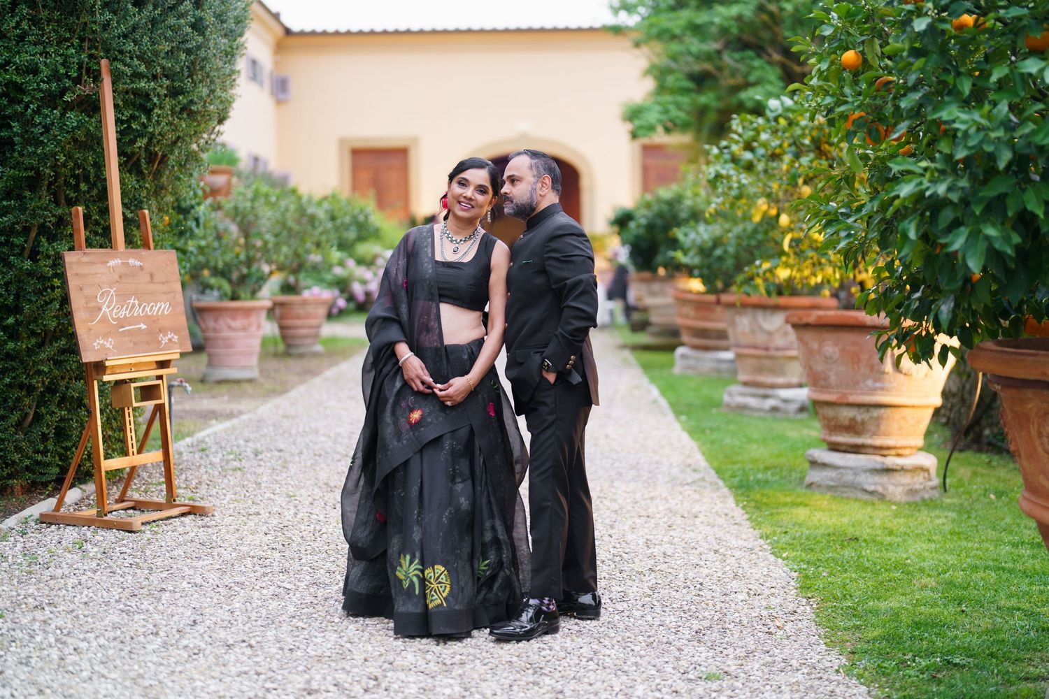 Couple in elegant black attire poses together on a gravel path lined with potted plants in a garden setting.