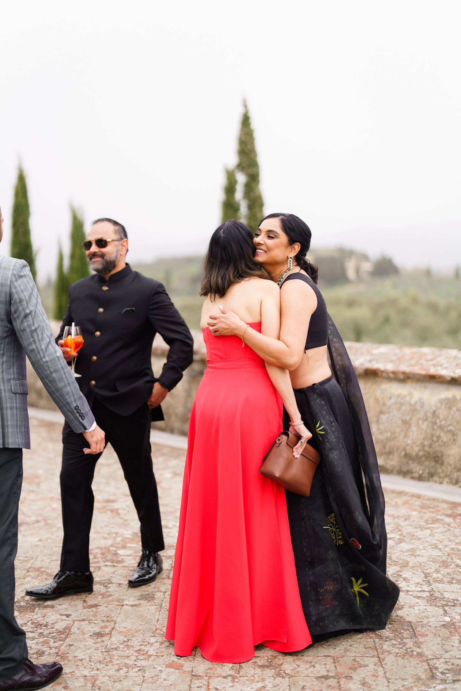 Group in formal attire gathers on stone terrace with cypress trees in background.