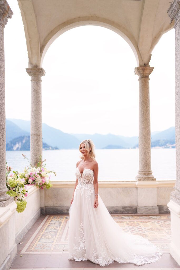 Elegant bridal portrait in white lace gown under Italian stone archway with lake view.