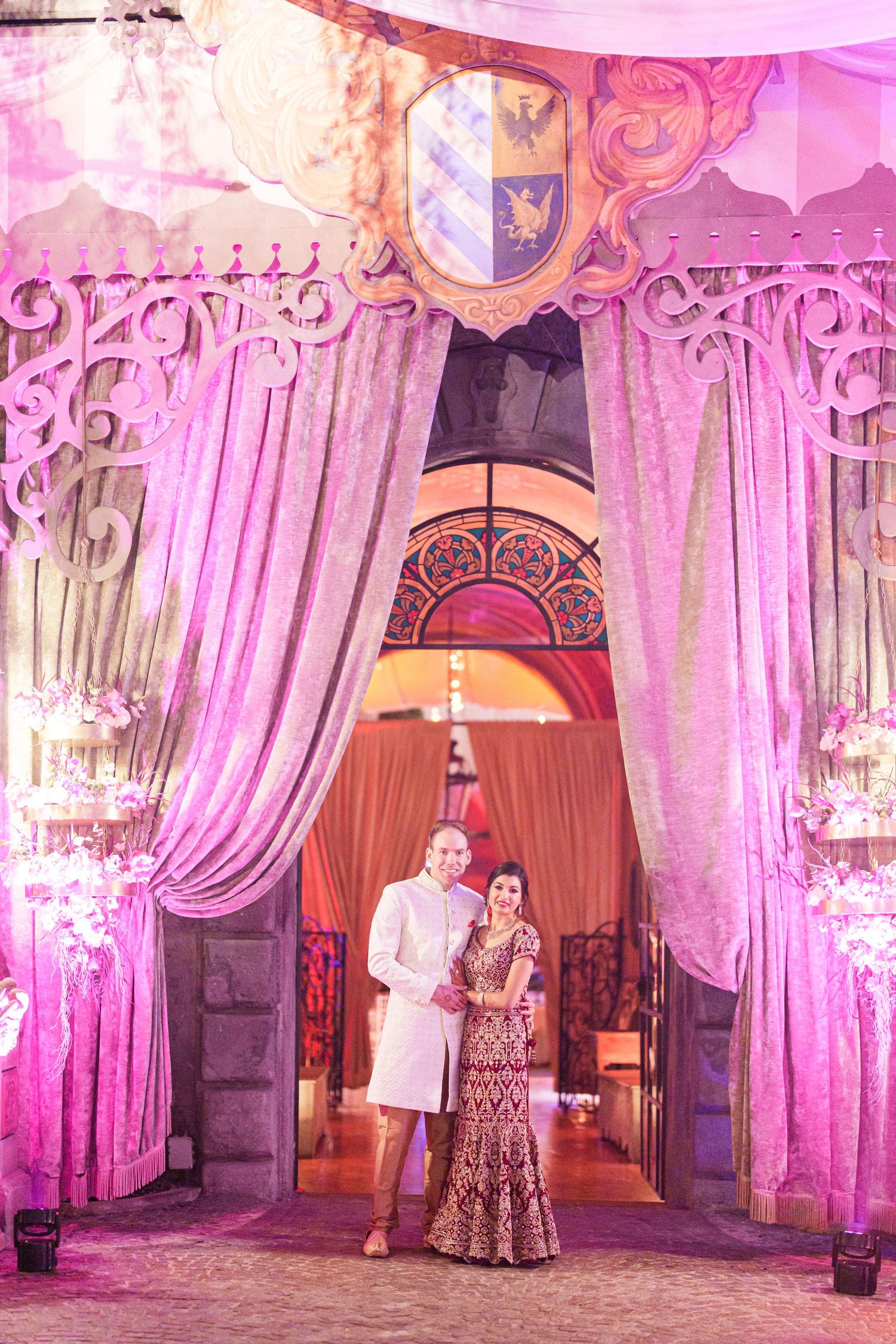 Elegant reception venue with pink uplighting, crystal chandeliers, and flowing fabric drapes.