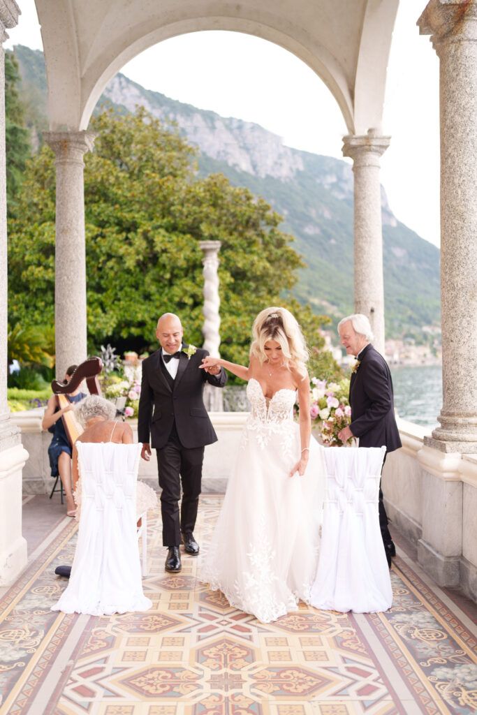 Wedding party walks through ornate columned terrace with mountain views and patterned tile floor.
