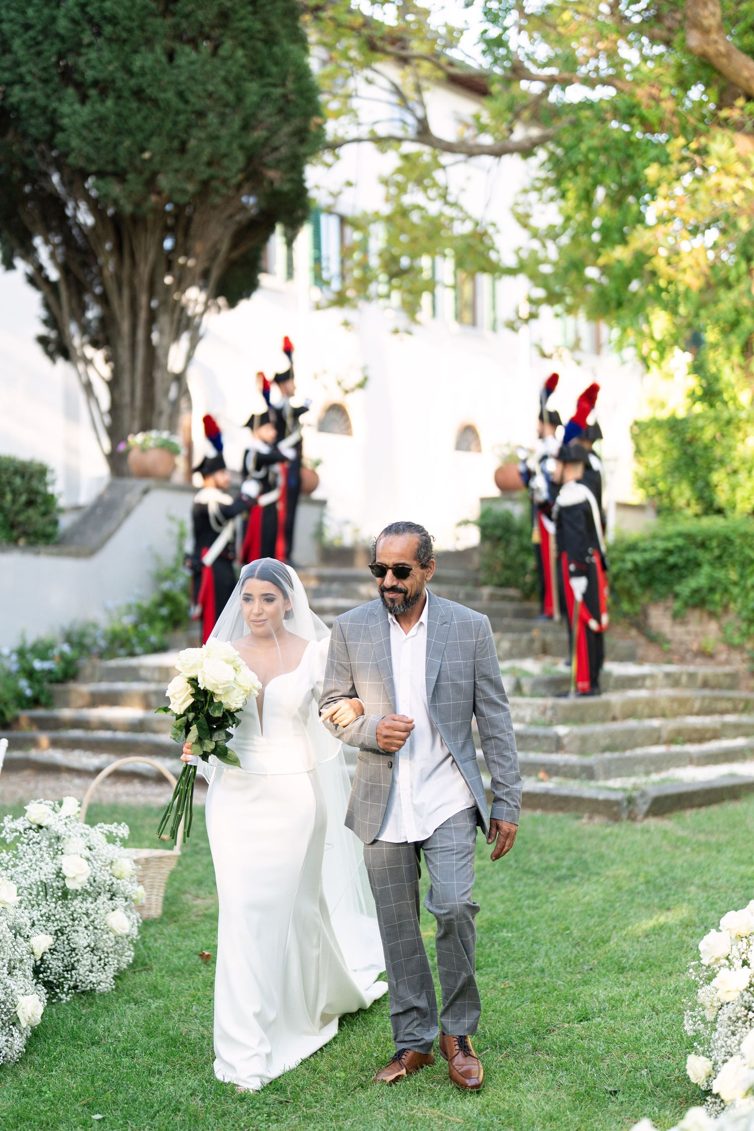 A couple walks along a garden path during an outdoor wedding ceremony with white floral decorations.