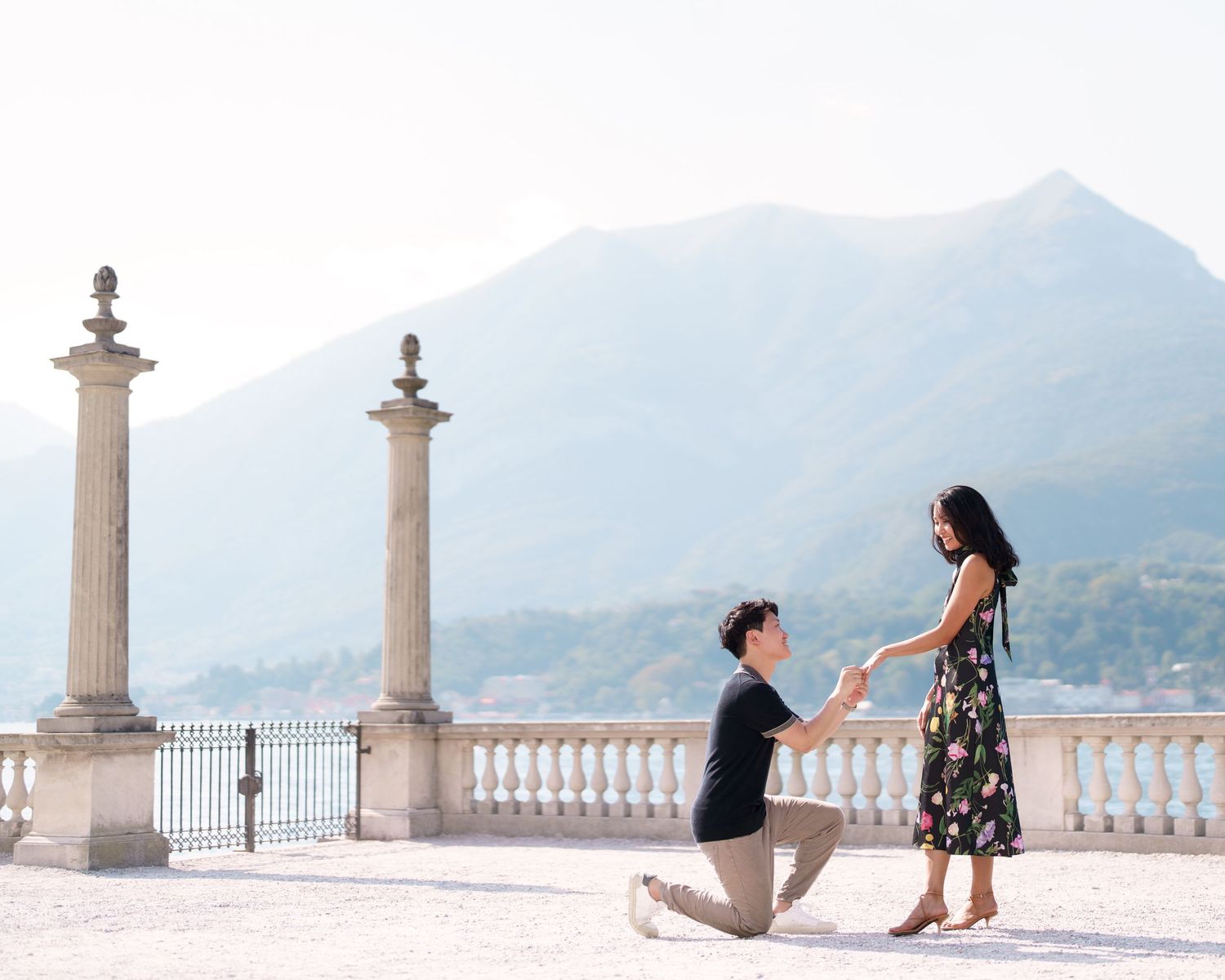 Two people sit and stretch on a white stone balustrade overlooking scenic mountains and water on a sunny day.