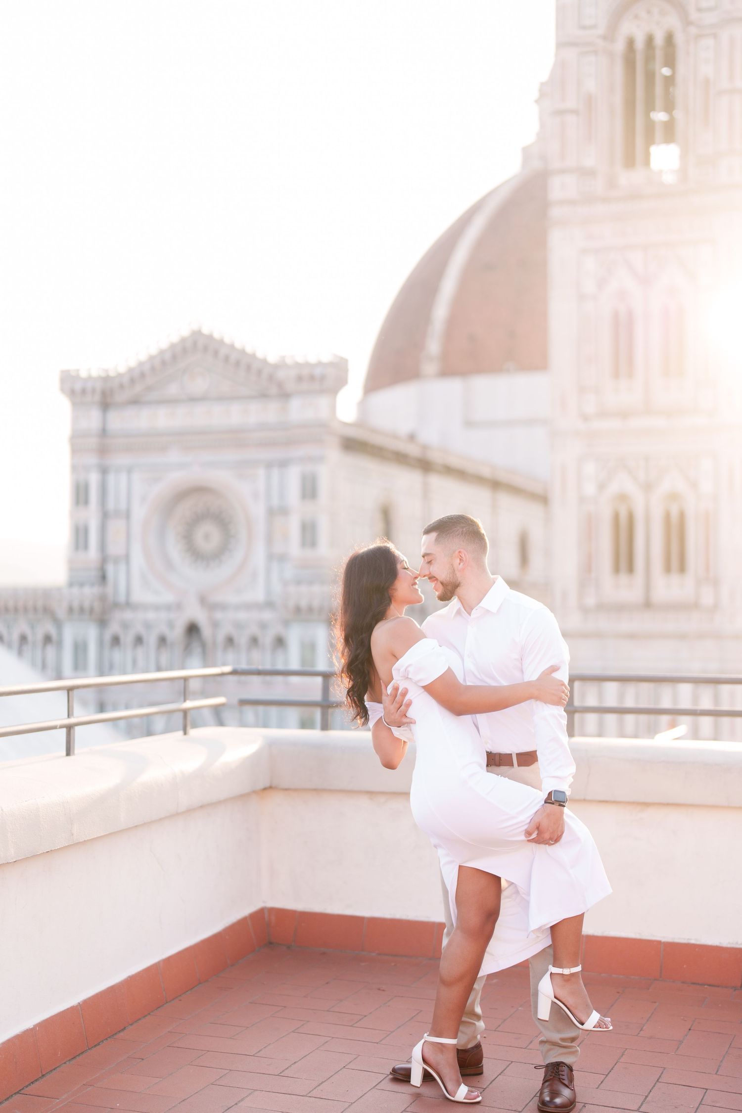 Romantic couple in white outfits embrace on a rooftop terrace overlooking Florence Cathedral during sunset in Italy.