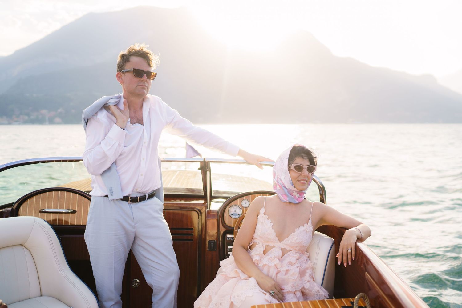 Couple on a luxury wooden boat on a serene lake with mountain backdrop, golden sunlight glowing behind them.