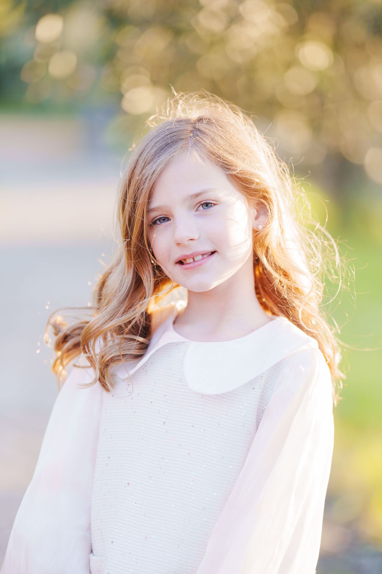 Young girl with long blonde hair smiling outdoors in a white sweater with bokeh background.