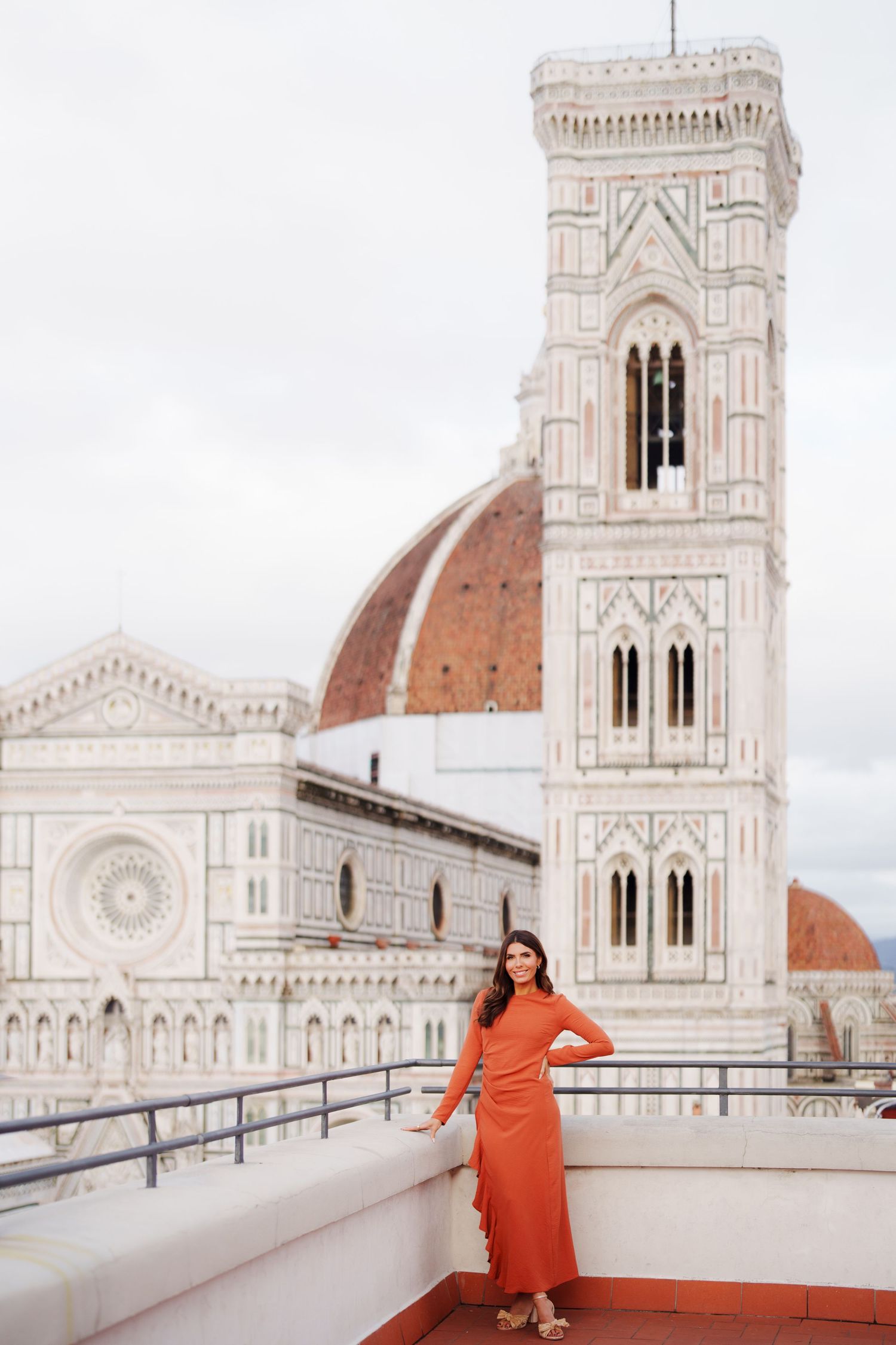 Woman in orange dress posing on rooftop terrace with Florence Cathedral and Giotto's Campanile in background.